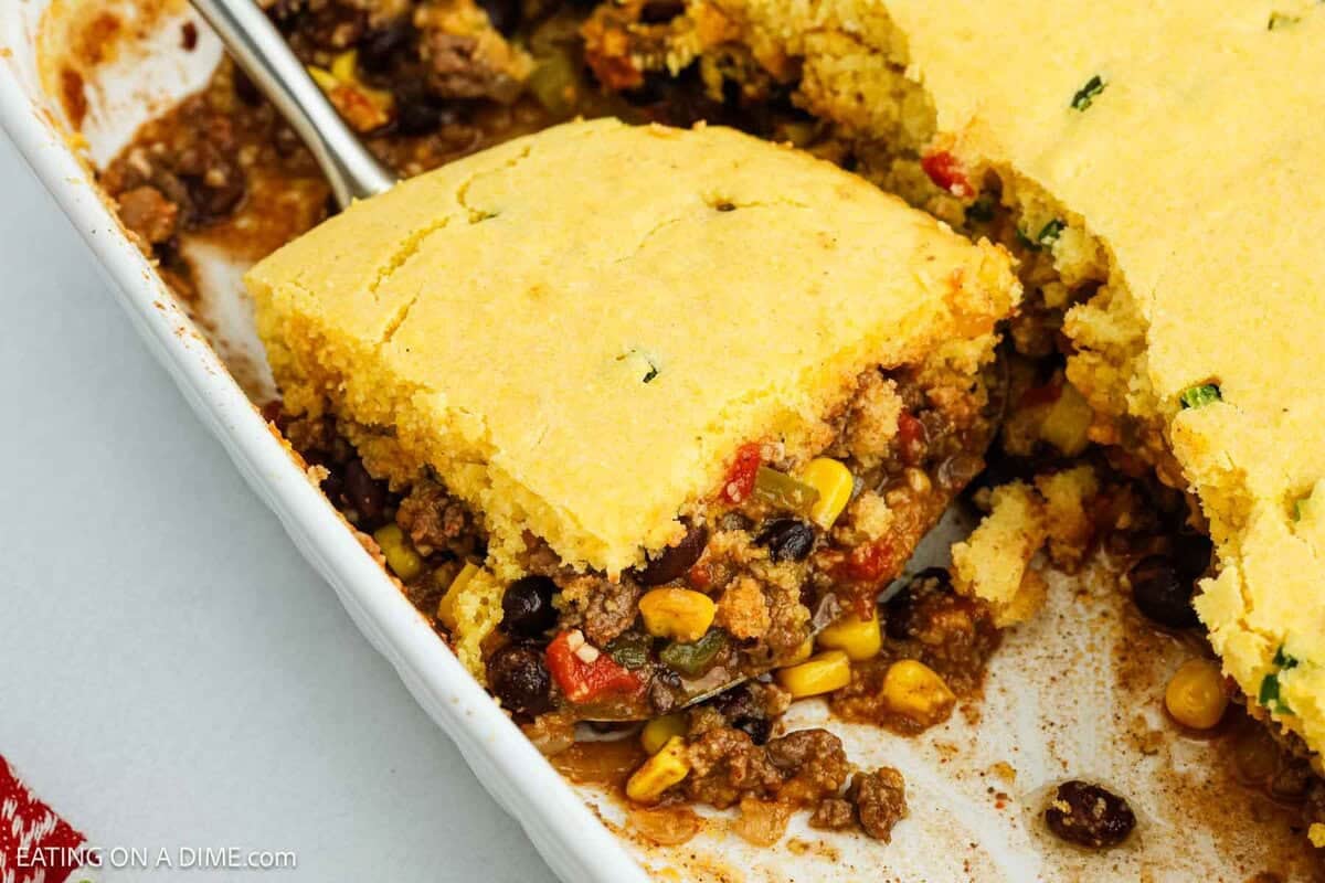 A baking dish filled with Mexican Cornbread Casserole, its golden cornbread topping revealing a hearty filling of ground beef, corn, black beans, and diced tomatoes. A serving is cut and partially lifted with a spatula.