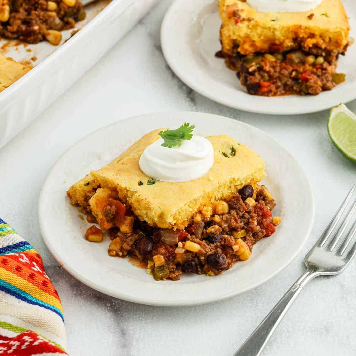 A slice of Mexican cornbread casserole with ground beef, black beans, corn, and veggies is served on a white plate with sour cream and cilantro, next to a fork and a colorful napkin.