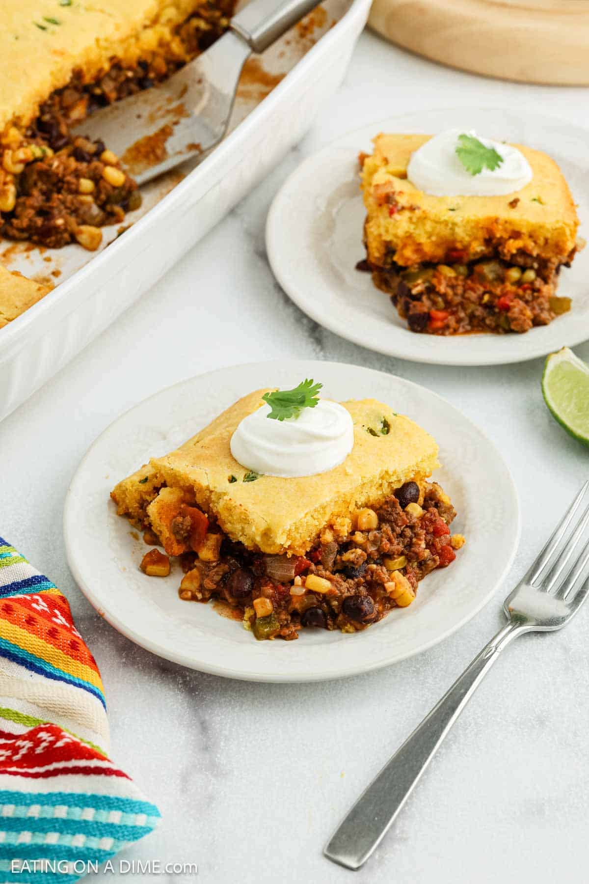 A serving of Mexican Cornbread Casserole with ground beef, beans, and vegetables is plated, topped with sour cream and a cilantro leaf. In the background sits a dish of more casserole, along with a colorful napkin and a fork.