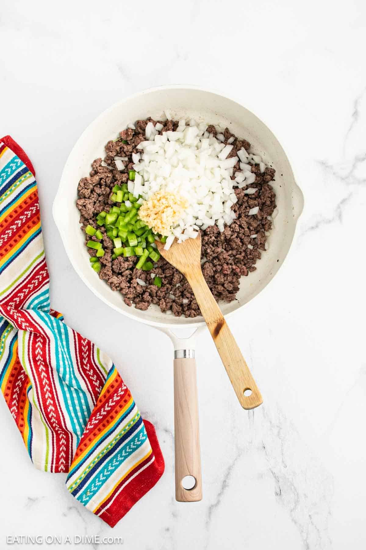 A white skillet with cooked ground beef, chopped onions, green bell peppers, and minced garlic is being stirred with a wooden spoon, the perfect start for a Mexican Cornbread Casserole. A colorful striped kitchen towel rests nearby on a white marble surface.