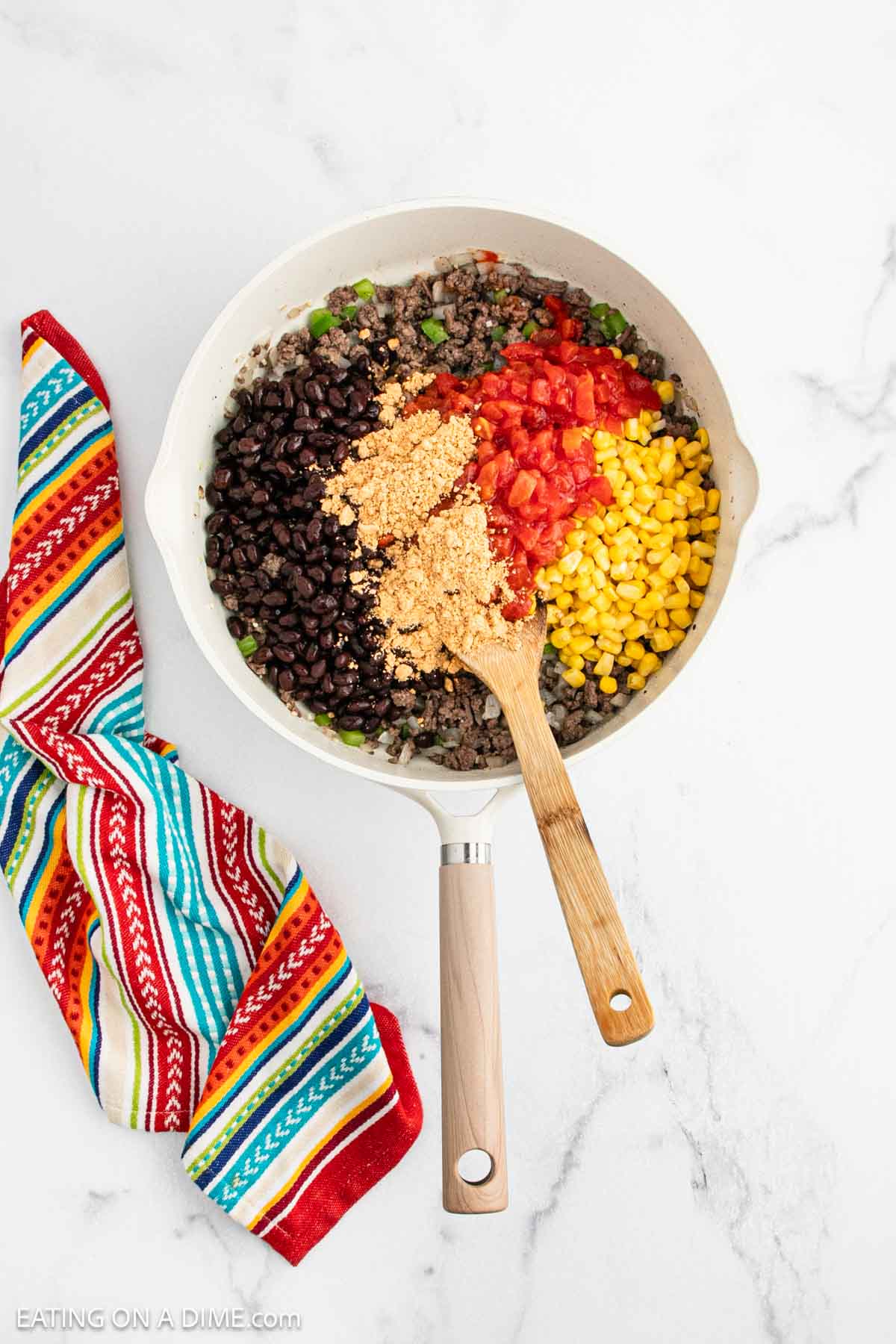 A white skillet filled with black beans, corn, diced tomatoes, cooked ground beef, and seasonings—perfect for a flavorful Mexican Cornbread Casserole—stirred with a wooden spoon. A colorful striped towel rests nearby on the marble surface.