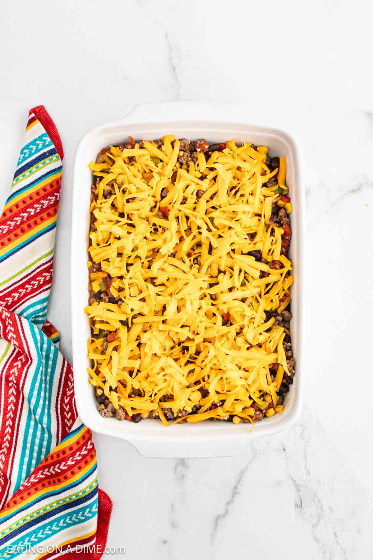 A white baking dish filled with Mexican Cornbread Casserole, topped with shredded cheddar cheese, sits on a marble surface next to a colorful striped cloth.