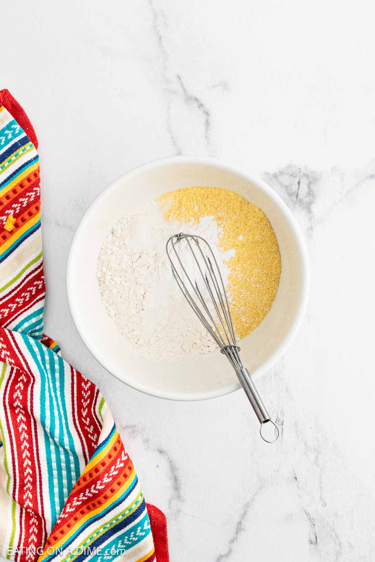 A white bowl with cornmeal, flour, and a whisk inside sits on a white marble surface next to a colorful striped kitchen towel, ready for mixing the base of a delicious Mexican Cornbread Casserole.