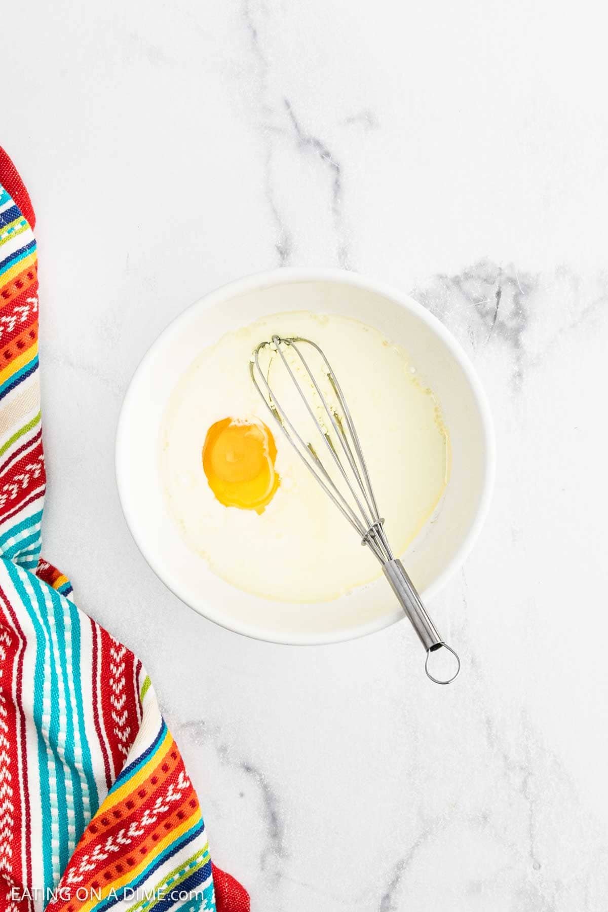 A white bowl with milk and a raw egg, partially whisked for cornbread casserole, sits on a white marble surface next to a colorful striped kitchen towel.