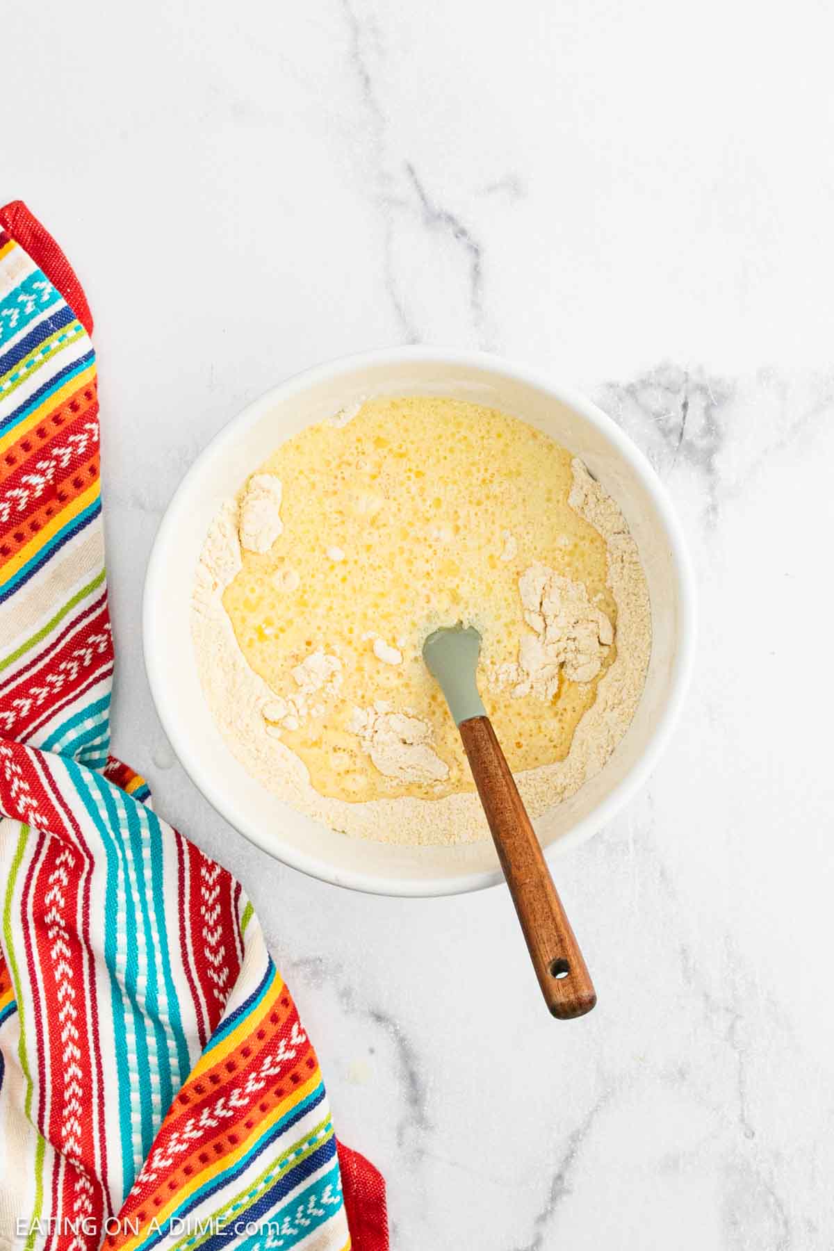 A white bowl with wet and dry ingredients partially mixed for Mexican cornbread casserole, and a spatula inside. The bowl is on a marble surface next to a colorful striped kitchen towel.