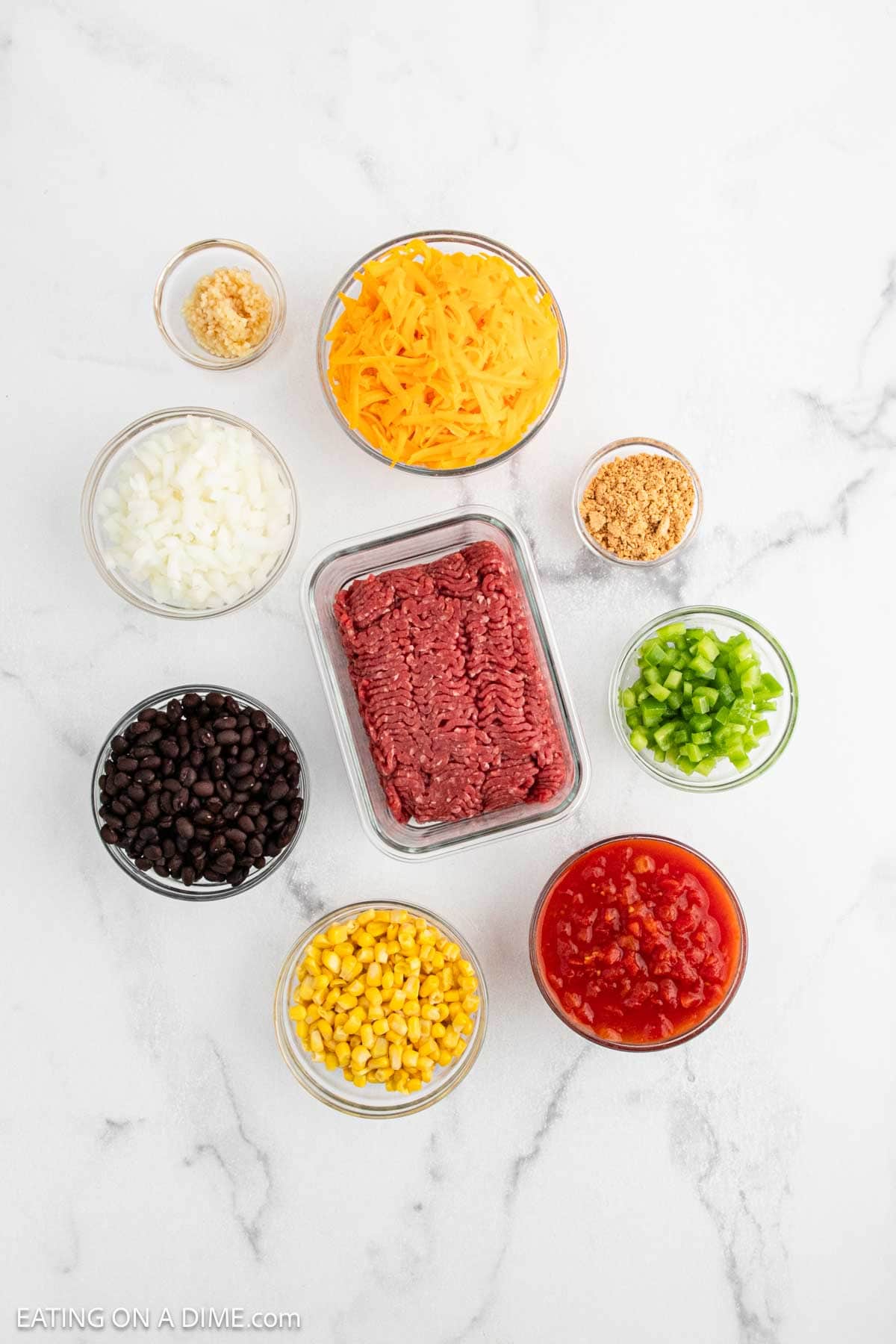Nine bowls on a white surface hold ingredients for a delicious Mexican Cornbread Casserole: shredded cheese, chopped onion, minced garlic, cooked ground beef, diced tomatoes, green bell pepper, crushed crackers, black beans, and corn.