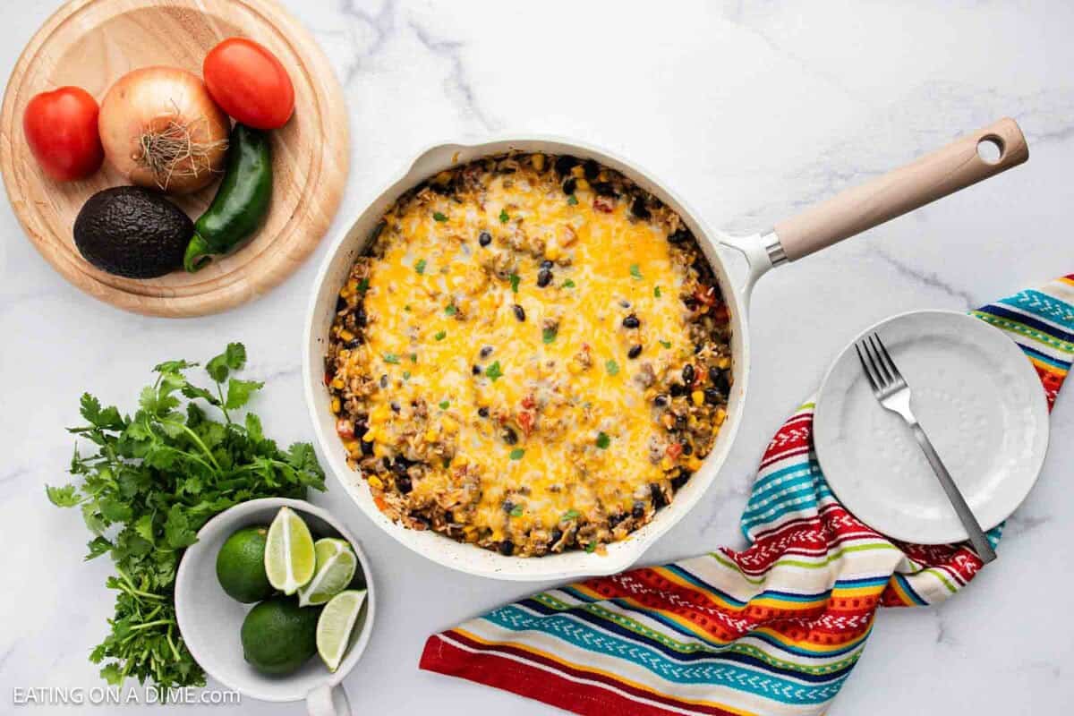 A skillet filled with a cheesy Mexican Rice Casserole sits on a white marble surface. Nearby are a colorful cloth, an empty plate with cutlery, and a cutting board with fresh vegetables and herbs, including tomatoes, onion, avocado, and lime.