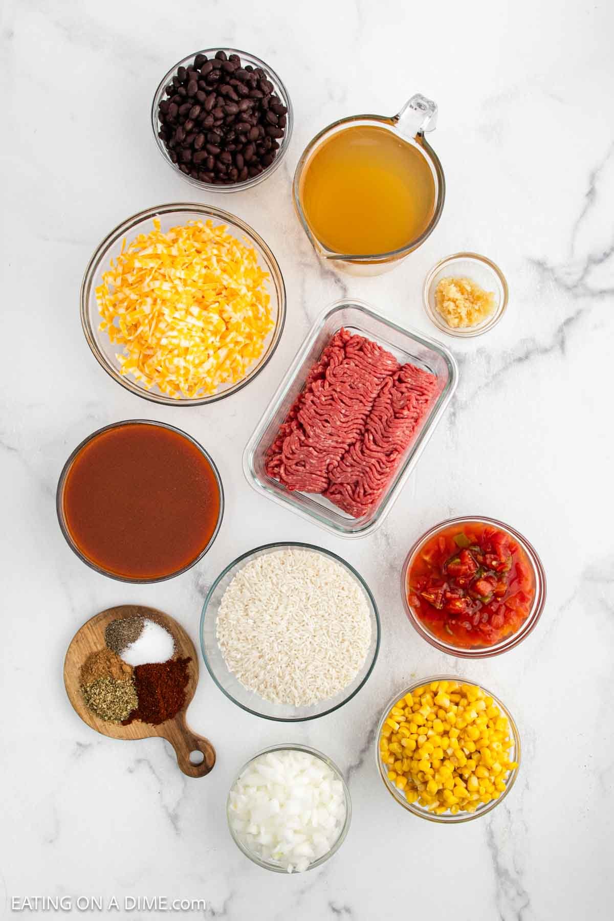 A top-down view of assorted ingredients for a Mexican Rice Casserole on a white surface, including ground beef, shredded cheese, rice, onions, corn, black beans, diced tomatoes, beef broth, minced garlic, chili sauce, and various spices.