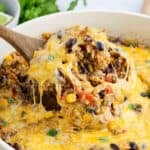 A wooden spoon lifts a cheesy, baked Mexican Rice Casserole of rice, ground beef, black beans, corn, and diced tomatoes from a skillet, with melted cheese stretching and a garnish of cilantro visible.