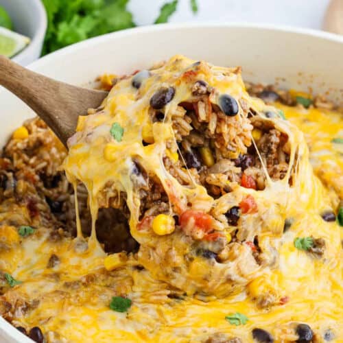 A wooden spoon lifts a cheesy, baked Mexican Rice Casserole of rice, ground beef, black beans, corn, and diced tomatoes from a skillet, with melted cheese stretching and a garnish of cilantro visible.