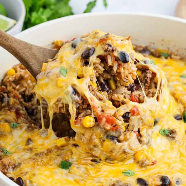A wooden spoon lifts a cheesy, baked Mexican Rice Casserole of rice, ground beef, black beans, corn, and diced tomatoes from a skillet, with melted cheese stretching and a garnish of cilantro visible.