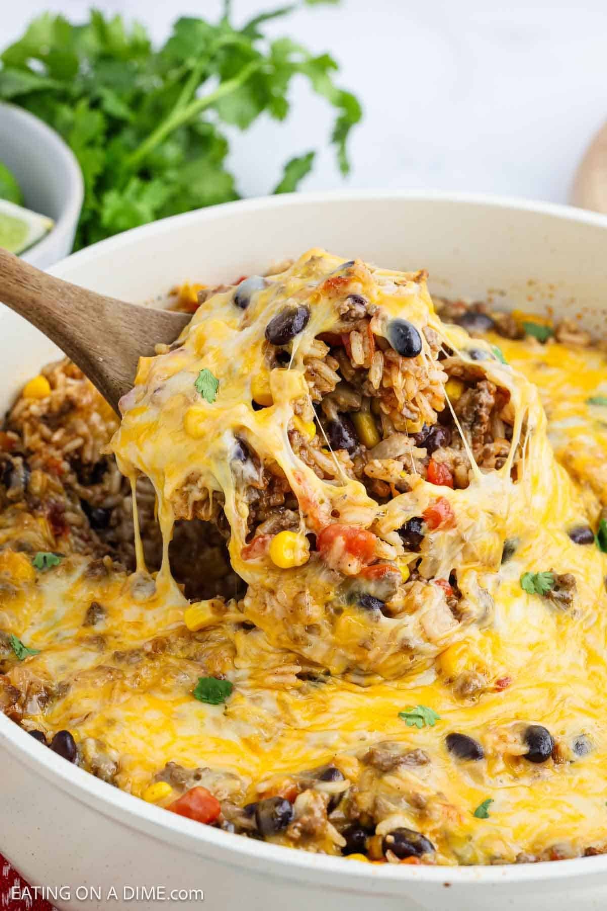 A wooden spoon lifts a cheesy Mexican Rice Casserole with black beans, corn, ground meat, and tomatoes from a white dish. Melted cheese stretches from the spoon, and fresh cilantro is visible in the background.