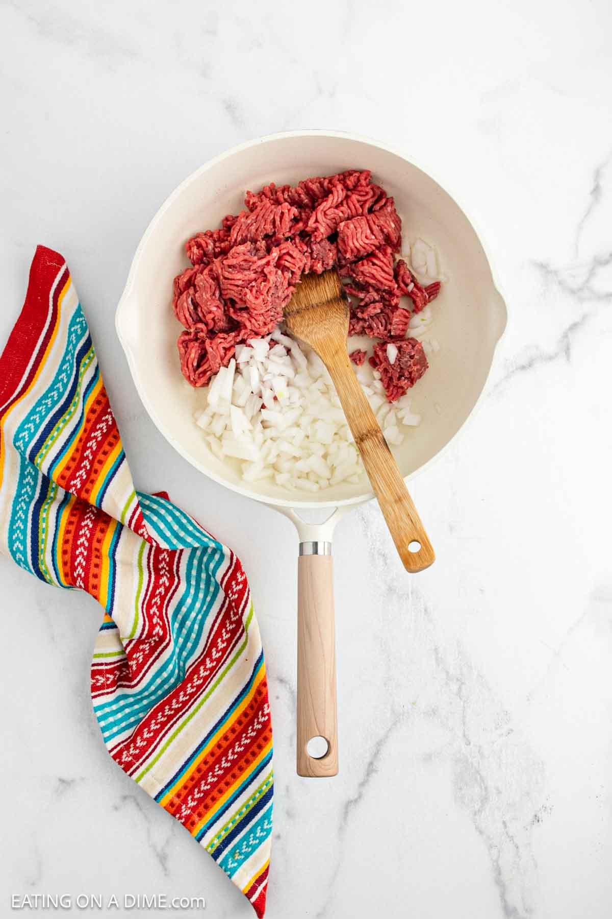 A white skillet with raw ground beef and chopped onions, partially mixed with a wooden spoon, is ready for a flavorful Mexican Rice Casserole. A colorful striped kitchen towel rests beside it on a white marble surface.
