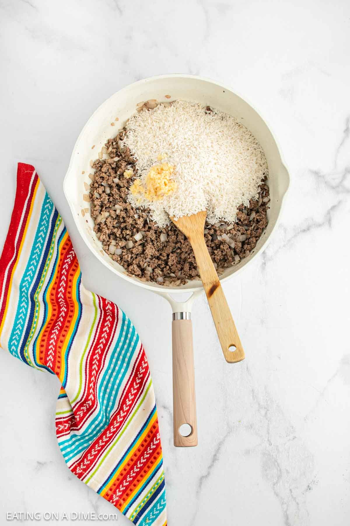 A white skillet on a marble surface holds Mexican Rice Casserole with cooked ground beef, onions, uncooked white rice, and grated cheese. A wooden spoon rests inside while a colorful striped kitchen towel lies beside the skillet.