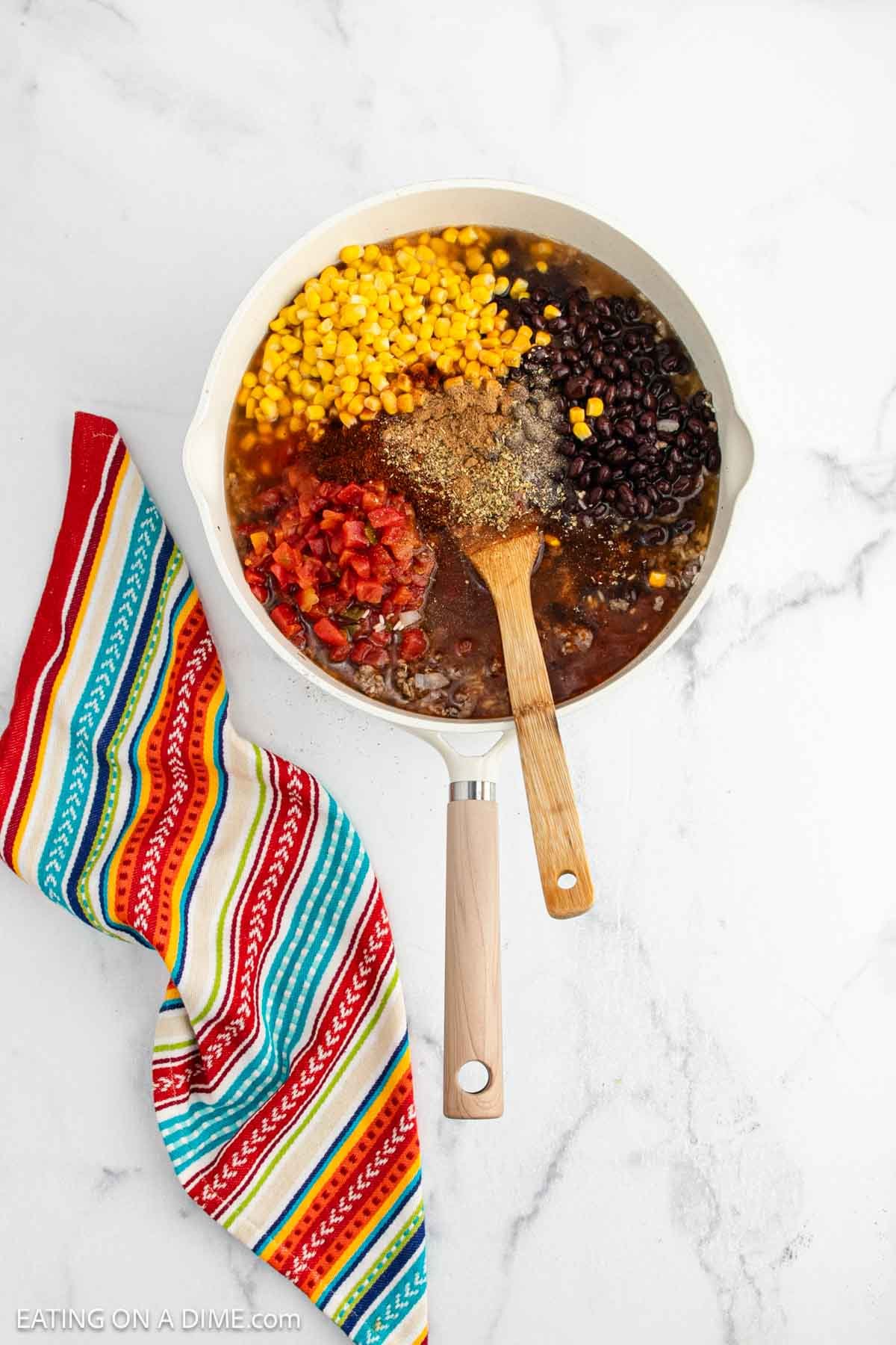 A white skillet with ground beef, corn, black beans, diced tomatoes, and spicesโperfect for a Mexican Rice Casseroleโsits with a wooden spoon. A colorful striped towel lies next to the skillet on a white marble surface.