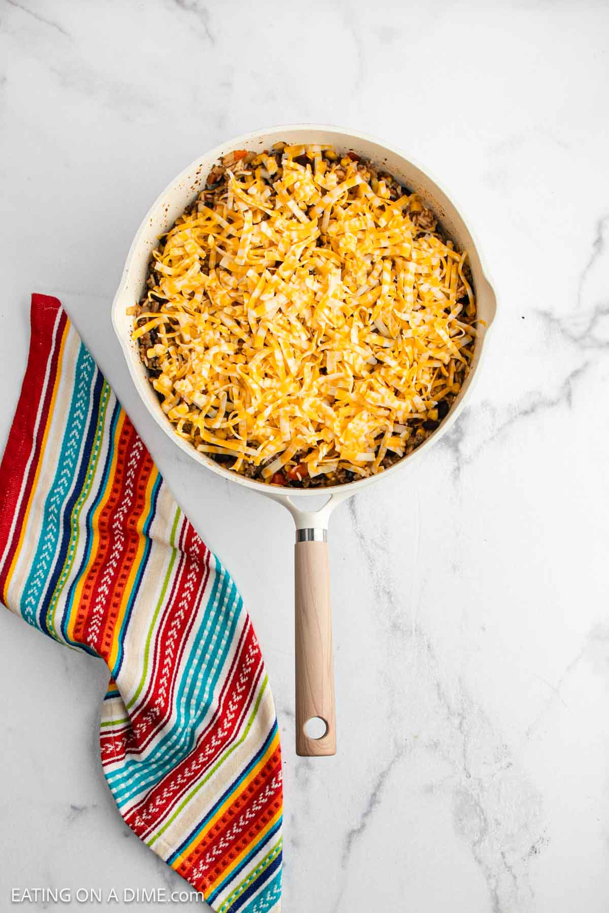 A skillet filled with a Mexican Casserole topped with shredded yellow and white cheese, next to a colorful striped cloth on a white marble surface.