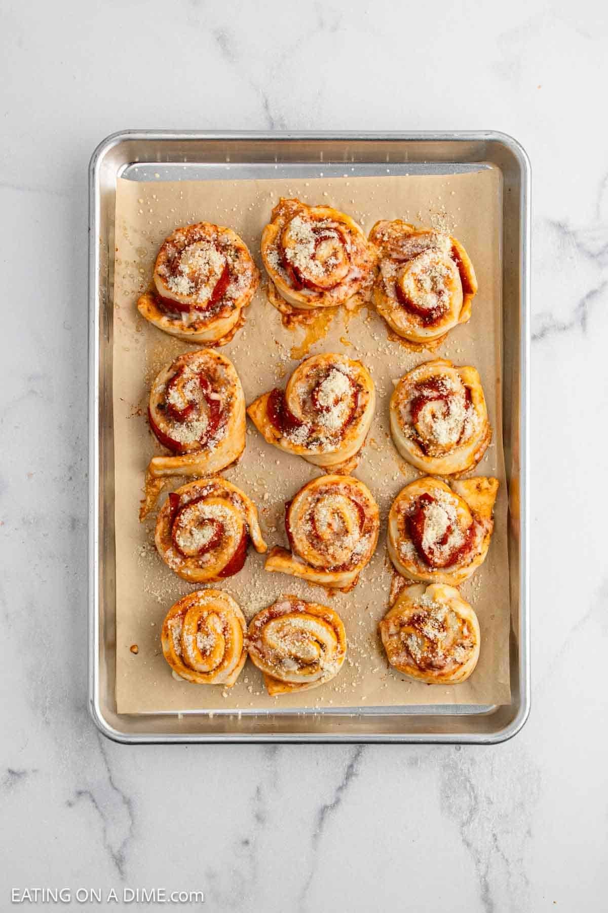 A baking sheet lined with parchment paper holds twelve baked pizza pinwheels topped with melted cheese and herbs, arranged in three rows on a marble countertop—perfect for trying out a new Pepperoni Roll recipe.