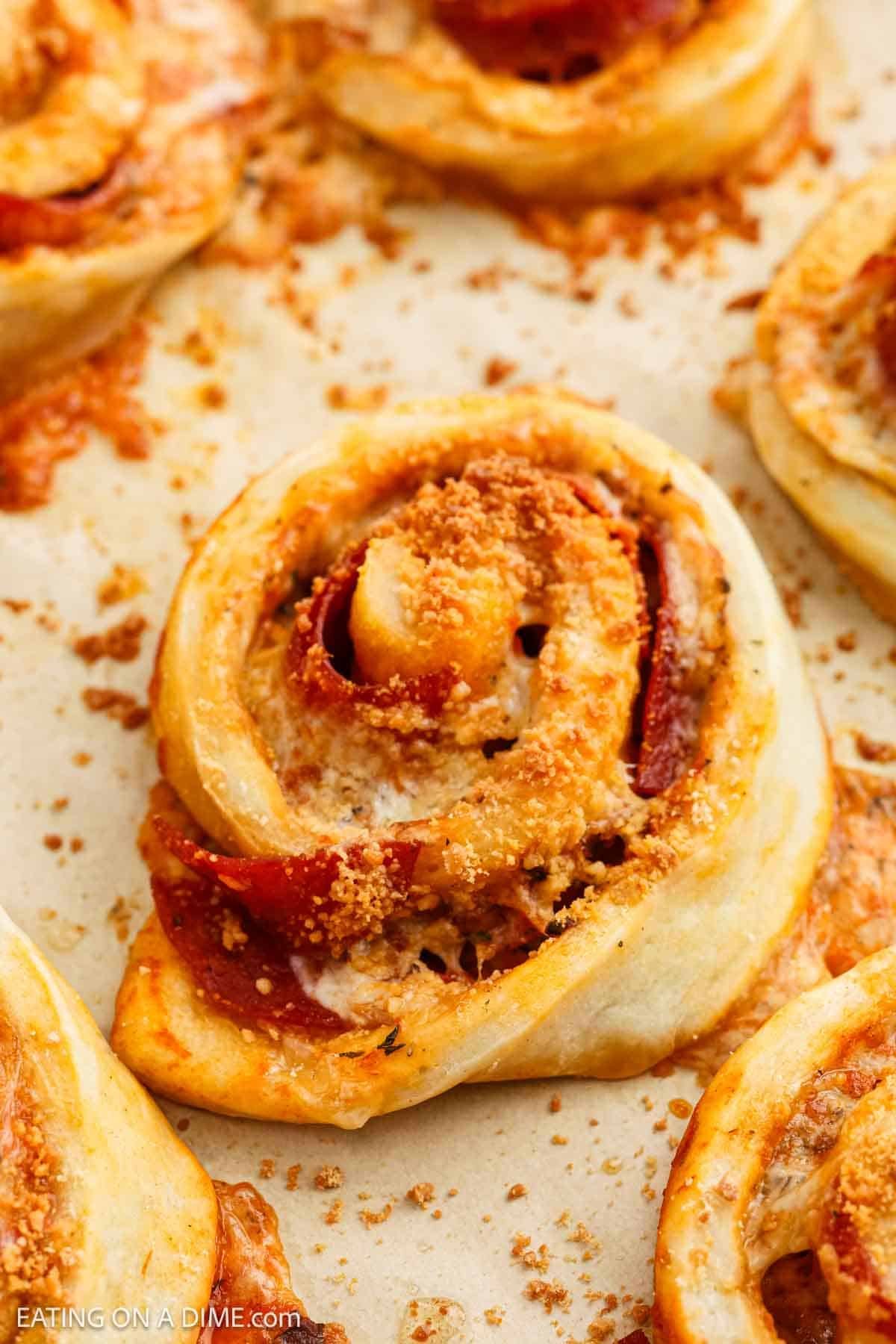 Close-up of baked pizza pinwheels, inspired by a classic Pepperoni Roll Recipe, showing golden-brown pastry swirled with melted cheese, pepperoni slices, and sprinkled with grated parmesan on a parchment-lined baking sheet.