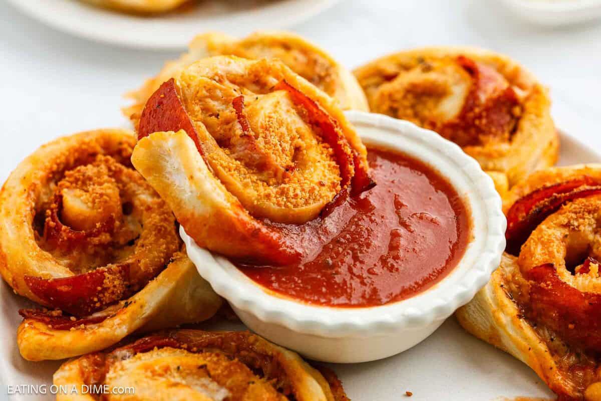 Close-up of golden brown pepperoni pizza rolls, made using a classic Pepperoni Roll Recipe, arranged around a white bowl of marinara sauce. One roll is being dipped, and all are topped with grated cheese.