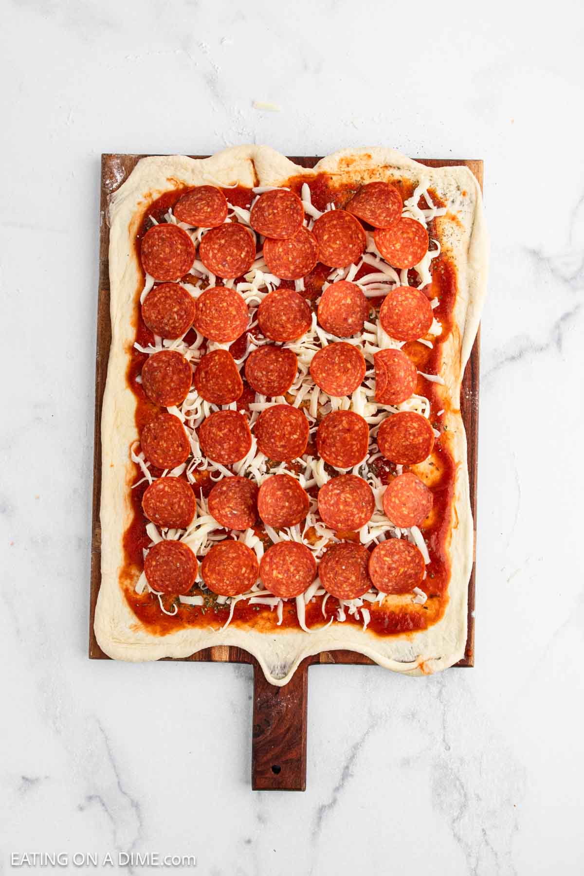 A rectangular uncooked pizza on a wooden board, topped with tomato sauce, shredded cheese, and evenly spaced pepperoni slices—perfect inspiration for your next Pepperoni Roll Recipe—rests on a white marble surface.