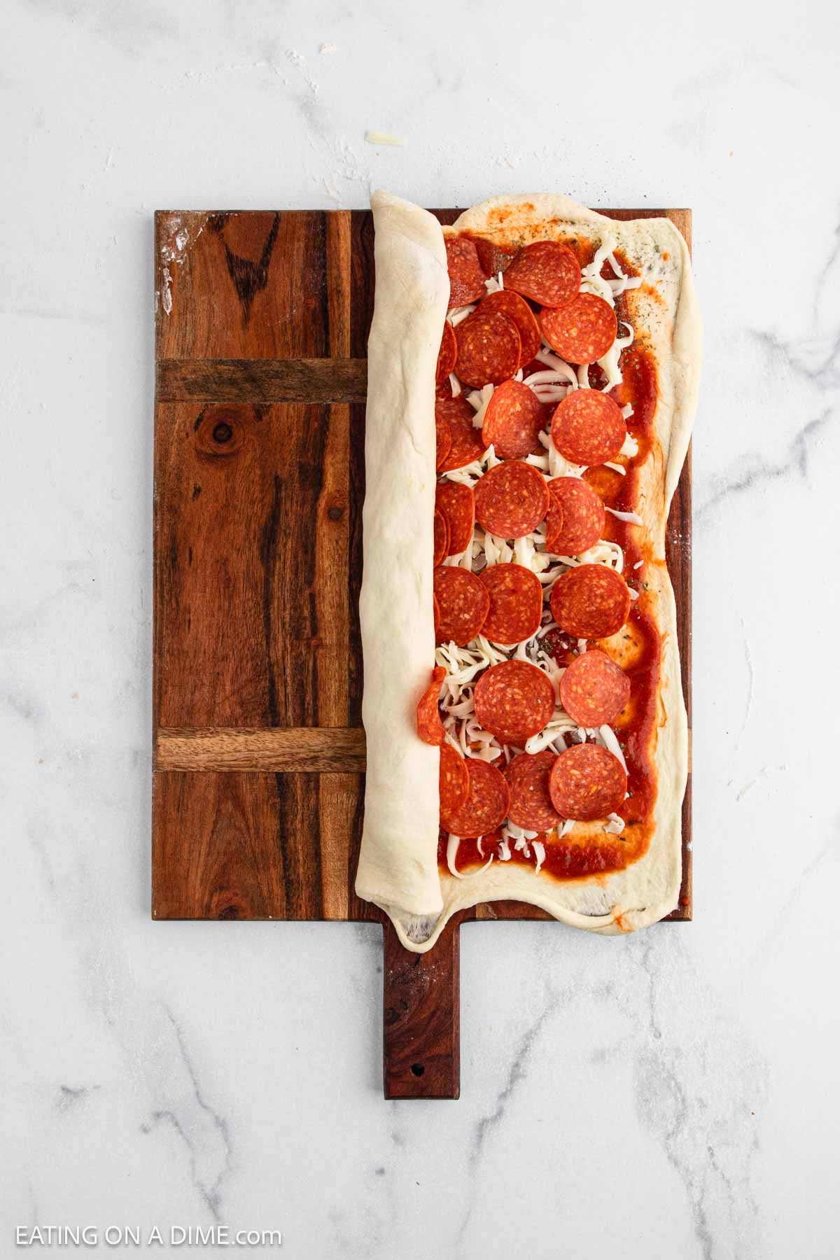 A wooden cutting board holds pizza dough topped with tomato sauce, shredded cheese, and pepperoni, partially rolled up—perfect for making a classic Pepperoni Roll. Try this easy Pepperoni Roll Recipe for a delicious homemade treat.