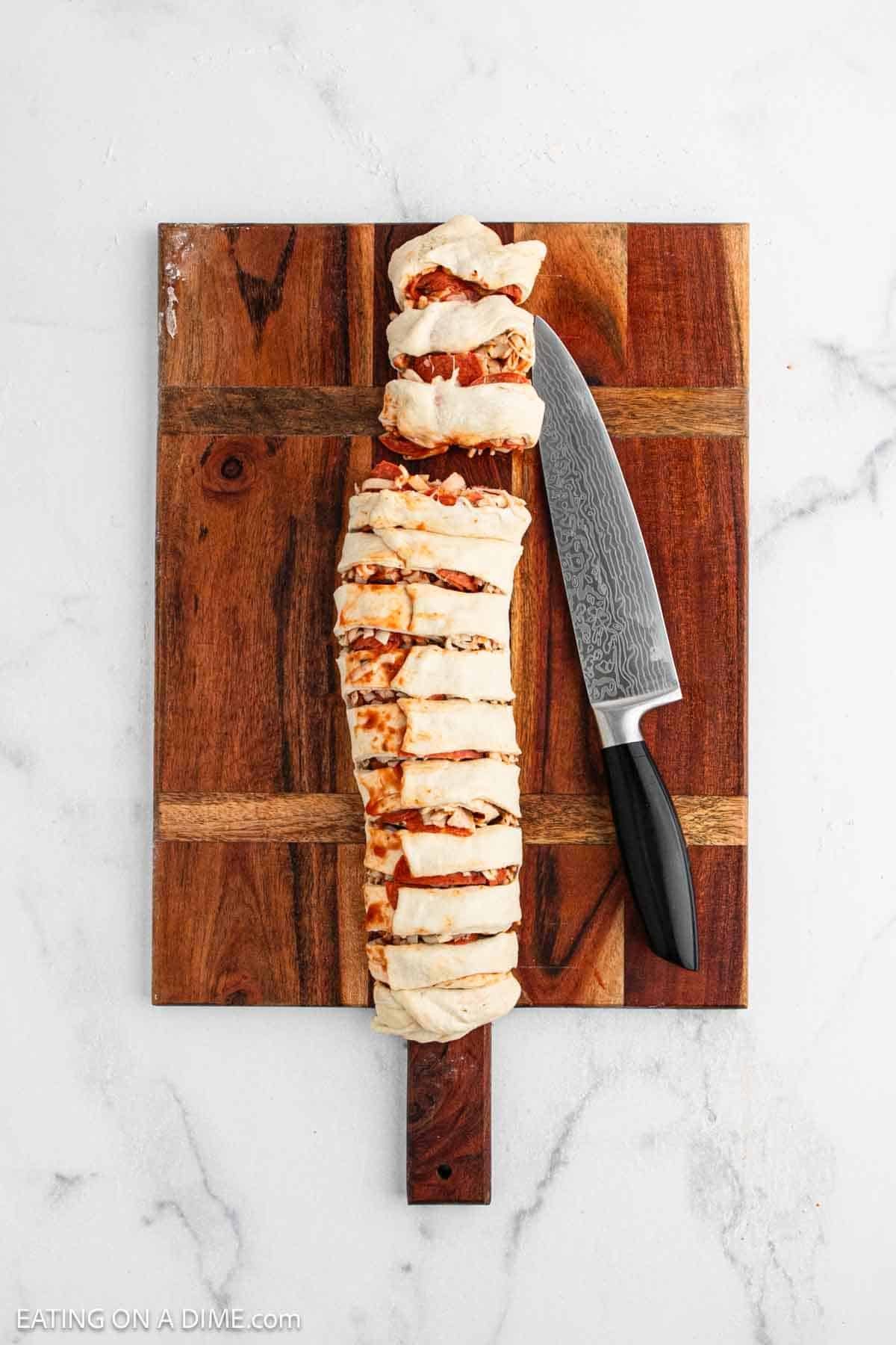 A sliced, baked homemade pepperoni roll sits on a wooden cutting board with a knife placed alongside it. The board rests on a white marble surface.