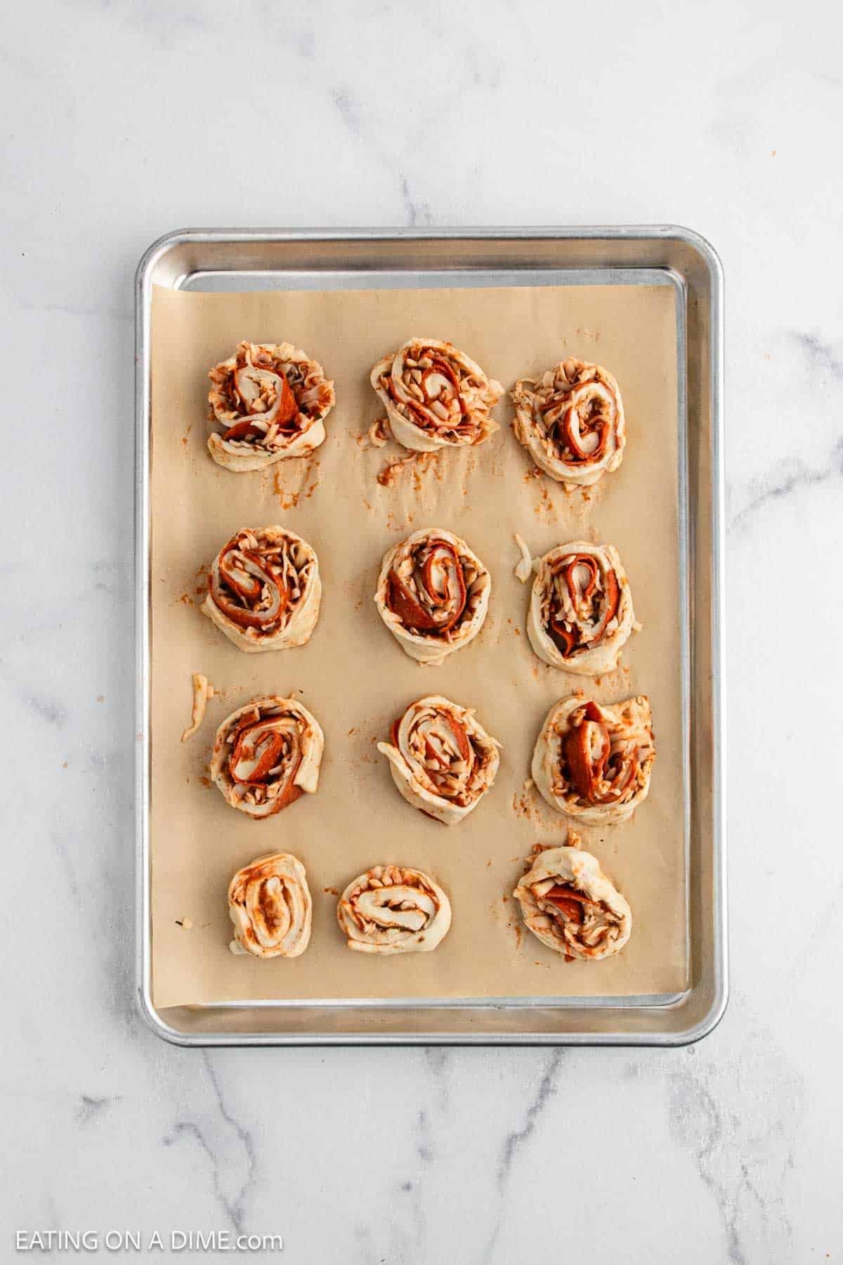 A baking sheet lined with parchment paper holds twelve uncooked pizza pinwheels, made from a classic Pepperoni Roll Recipe with dough, tomato sauce, and pepperoni, arranged neatly on a white marble surface.