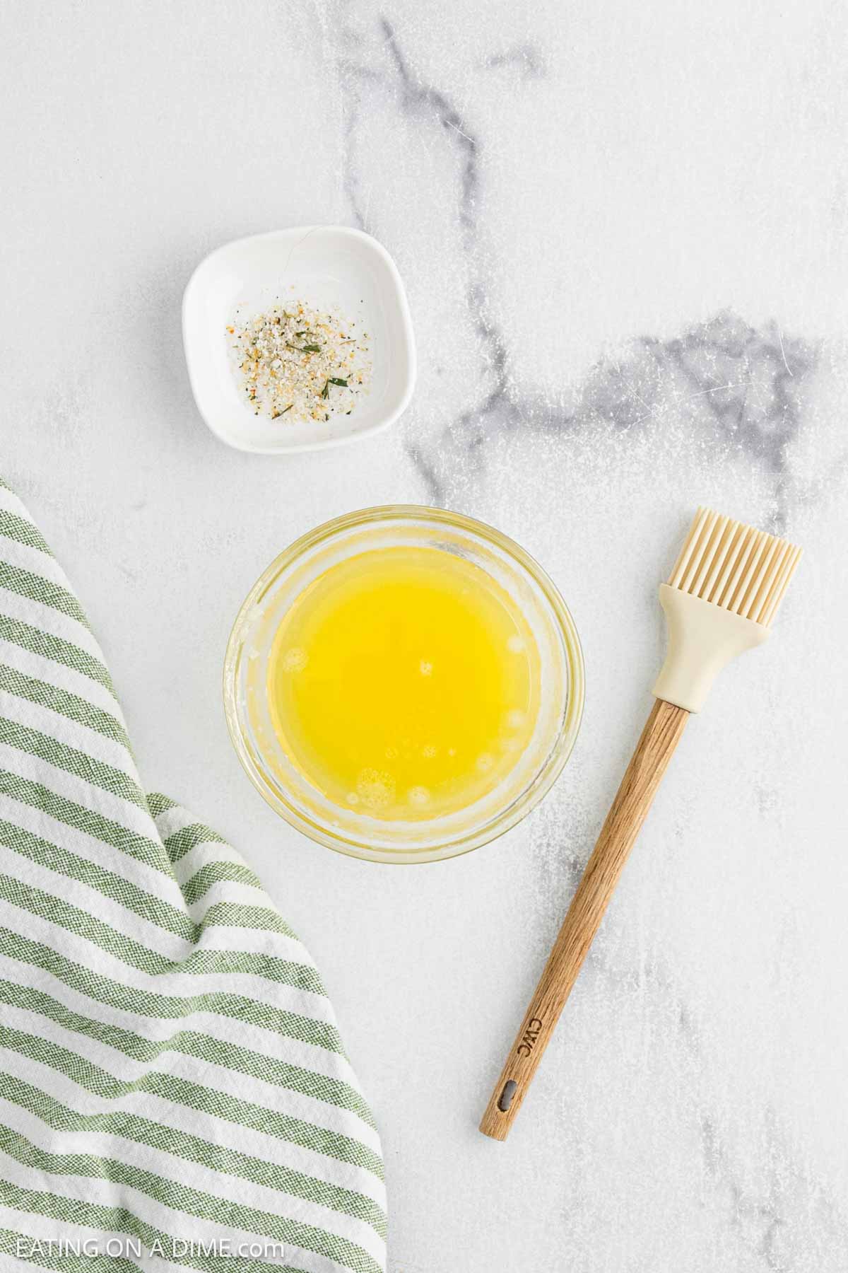 Overhead view of melted butter, seasoning, a pastry brush, and a green and white striped cloth on marble—perfect ingredients for starting a delicious Pepperoni Roll Recipe.