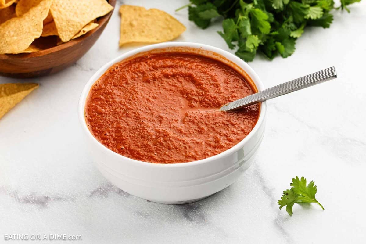 A white bowl filled with red ranchero sauce and a spoon, surrounded by tortilla chips and fresh cilantro on a white surface.