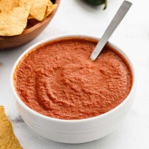 A white bowl filled with red ranchero sauce and a spoon, placed on a white surface. Tortilla chips are visible nearby on the left.