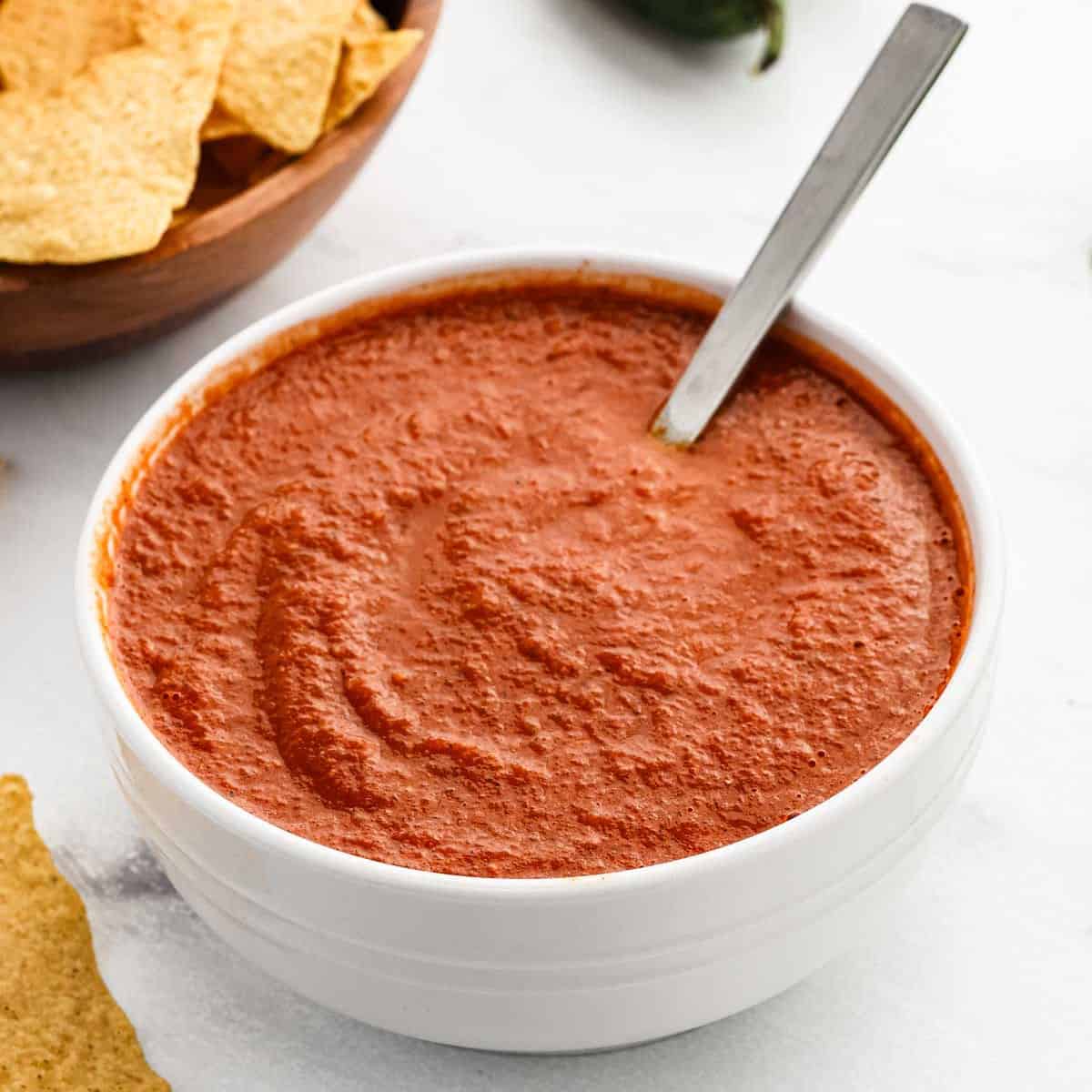 A white bowl filled with red ranchero sauce and a spoon, placed on a white surface. Tortilla chips are visible nearby on the left.