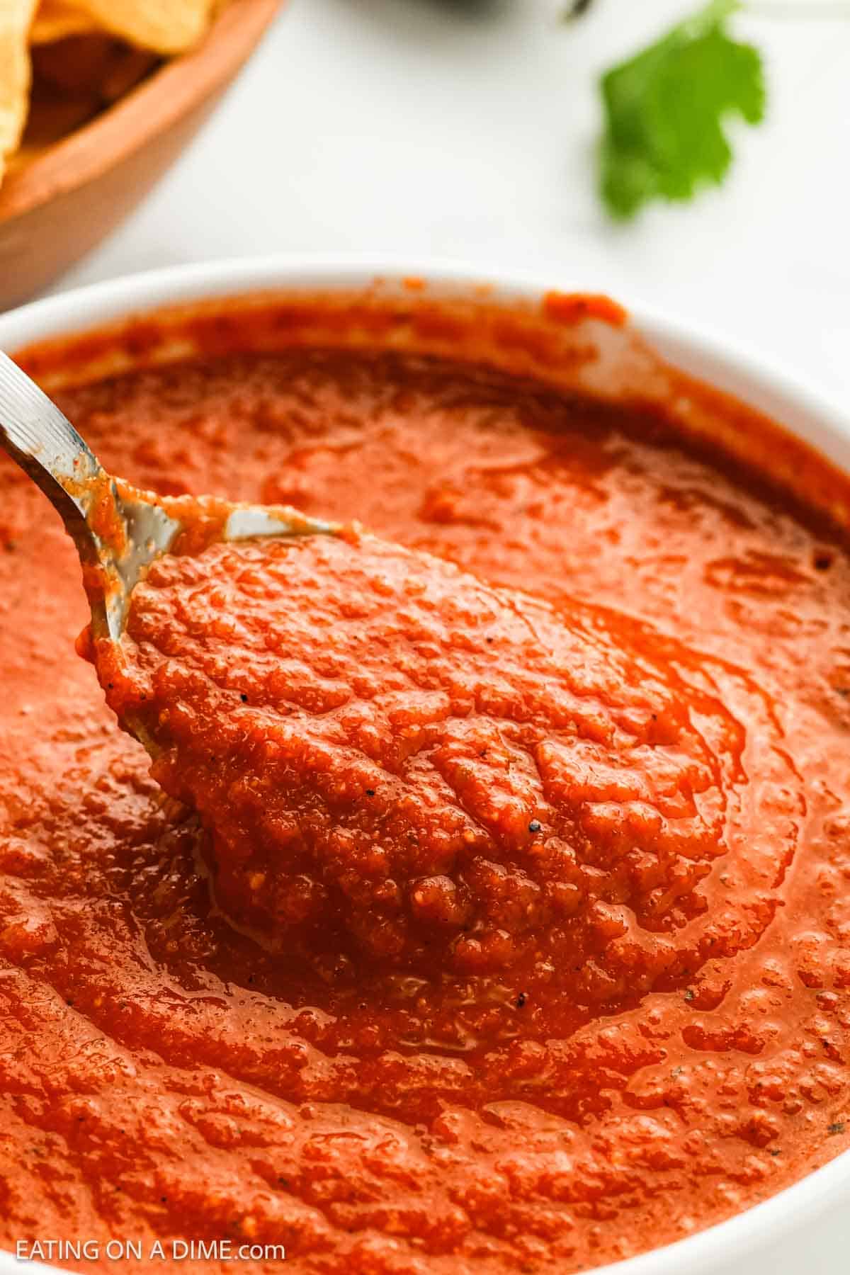 A close-up of a spoon lifting chunky Mexican sauce from a white bowl, with tortilla chips and a sprig of cilantro blurred in the background.