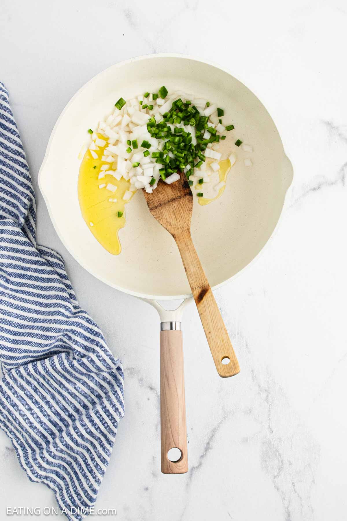 A white skillet with chopped onions and green peppers being sautรฉed in oil for ranchero sauce, stirred with a wooden spatula. A blue and white striped towel is next to the skillet on a marble countertop.