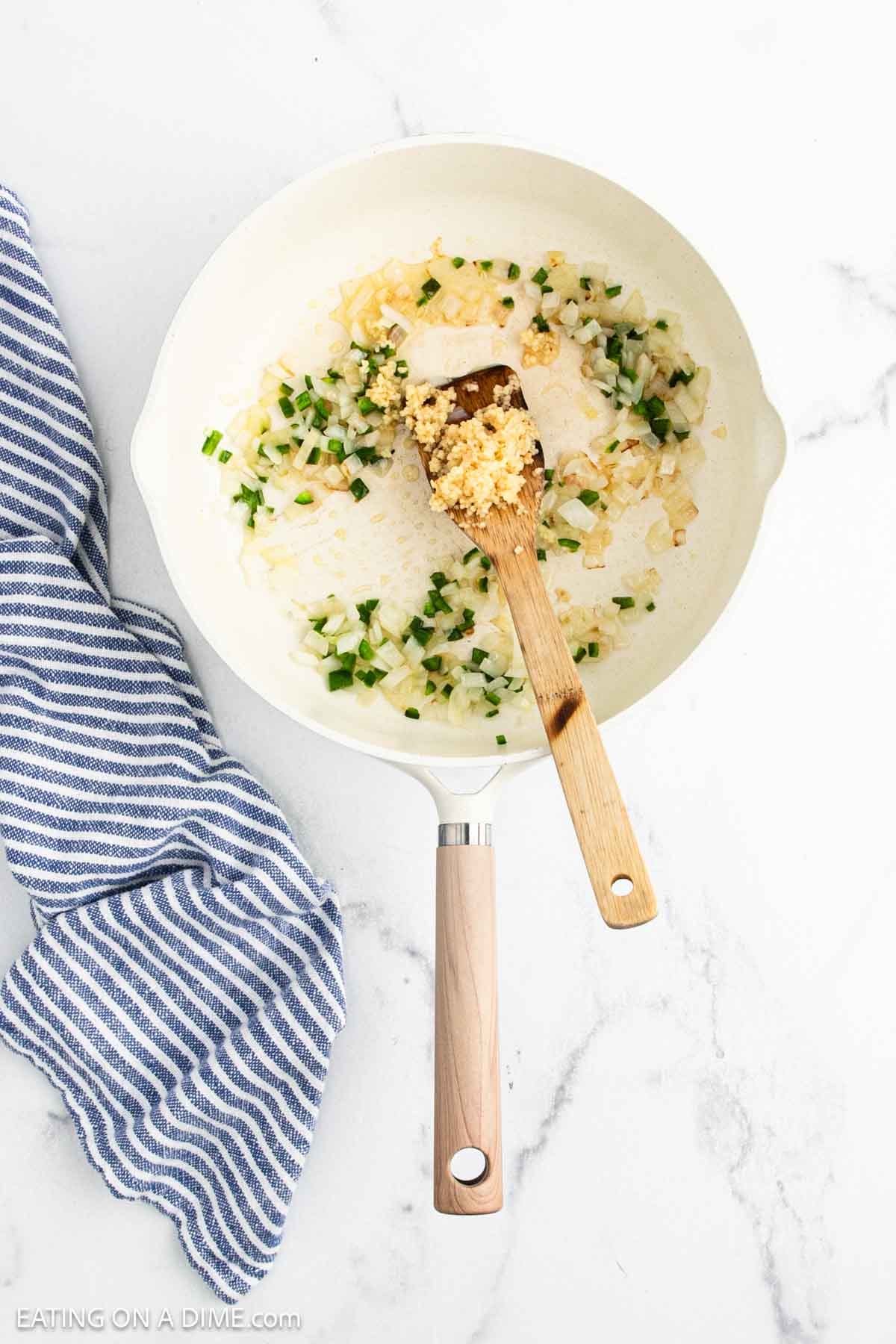 A white skillet holds chopped onions, green peppers, and minced garlic being sautรฉed for Ranchero Sauce with a wooden spatula. A blue and white striped kitchen towel rests beside the skillet on a marble countertop.