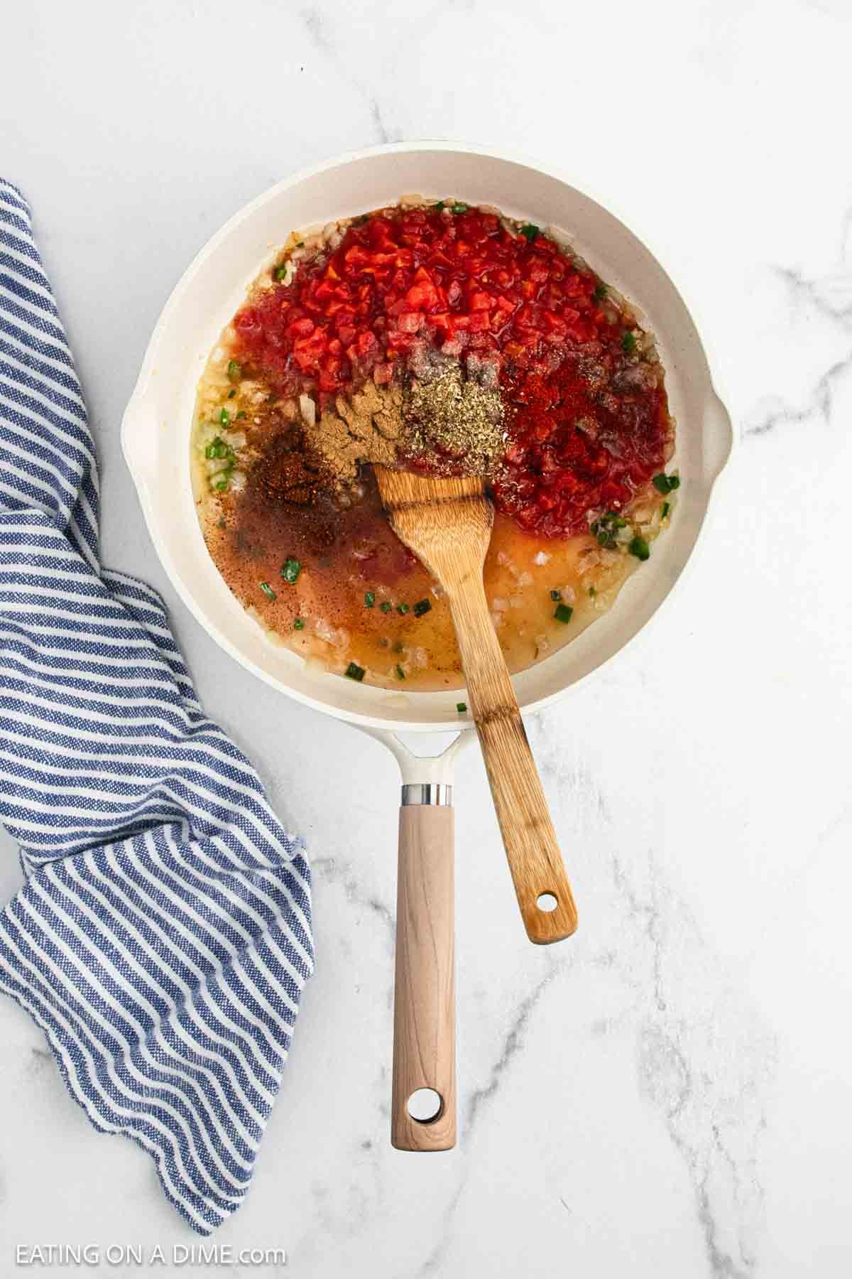 A skillet with chopped onions, red bell peppers, and assorted spices is being stirred into a vibrant Ranchero Sauce with a wooden spatula, next to a blue and white striped kitchen towel on a marble countertop.