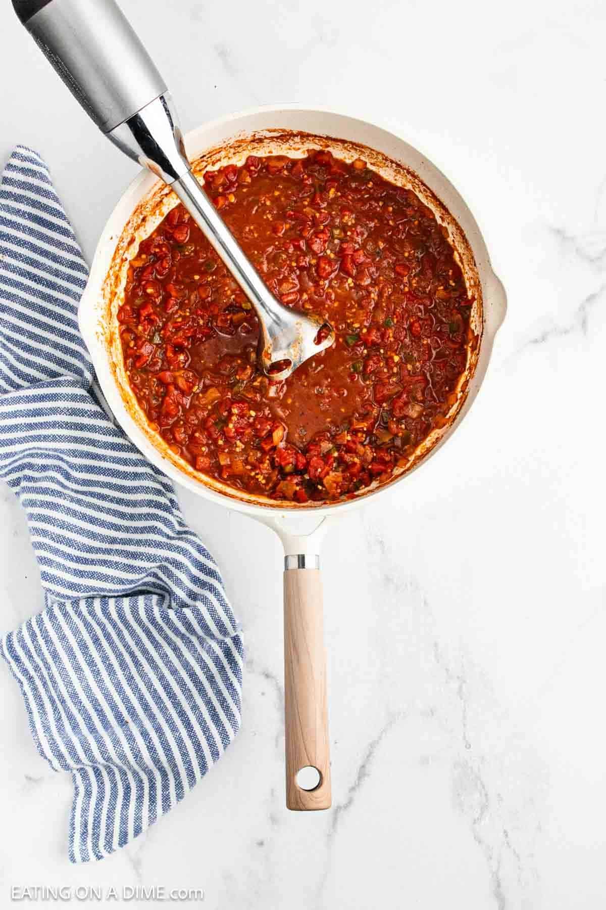 A white skillet filled with chunky Ranchero Sauce being blended with an immersion blender, next to a blue and white striped kitchen towel on a marble surface.