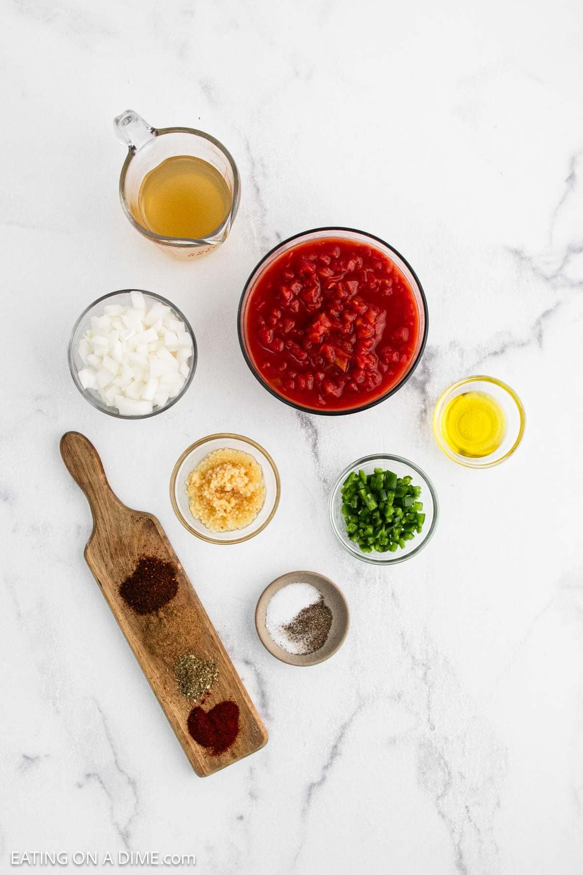 Top-down view of Ranchero Sauce ingredients on a white surface: chopped onions, minced garlic, chopped jalapeรฑos, olive oil, diced tomatoes, vegetable broth, and spices including chili powder, cumin, paprika, salt, and pepper.