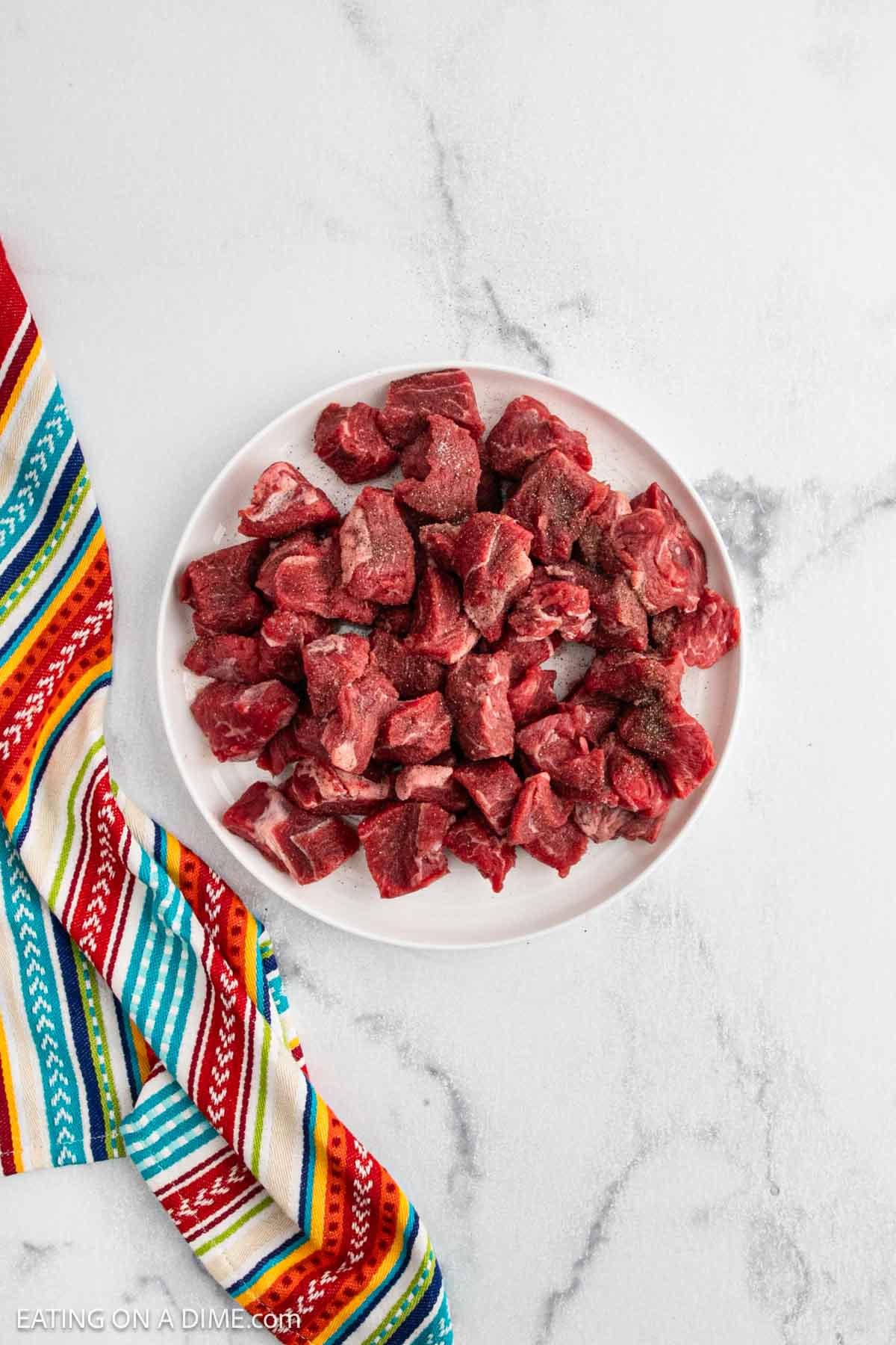 A white plate filled with raw, cubed beef sits on a light marble surface next to a colorful striped cloth.