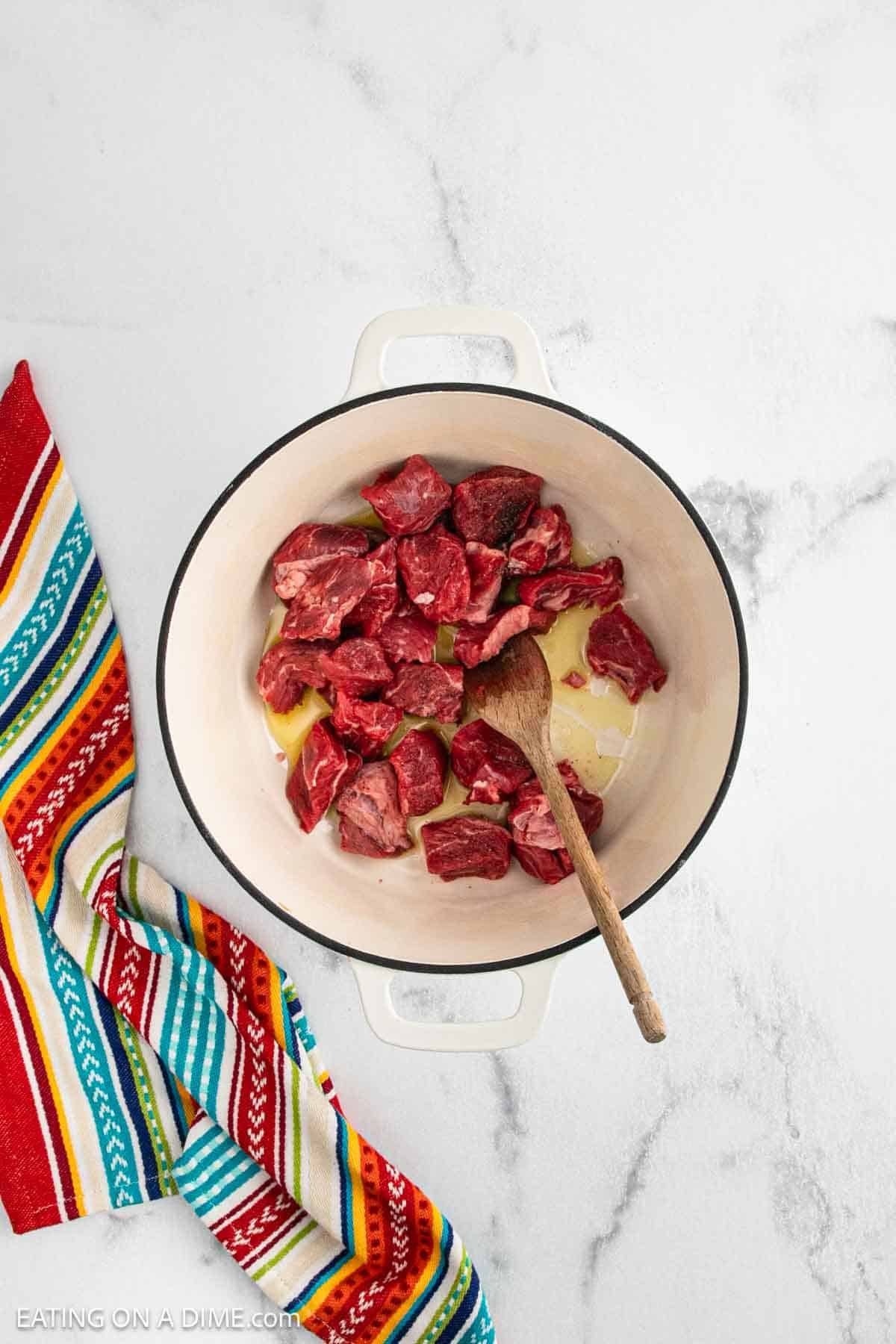 Overhead view of a white pot with raw beef chunks being browned in oil, stirred with a wooden spoon. A colorful striped kitchen towel is next to the pot on a white marble countertop.