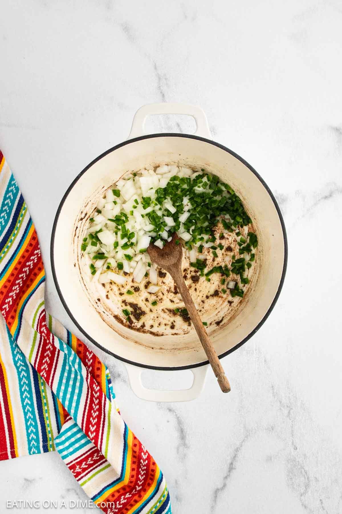 A white pot on a marble surface contains chopped onions and green peppers being sautรฉed, with a wooden spoon inside. A colorful striped cloth lies next to the pot.