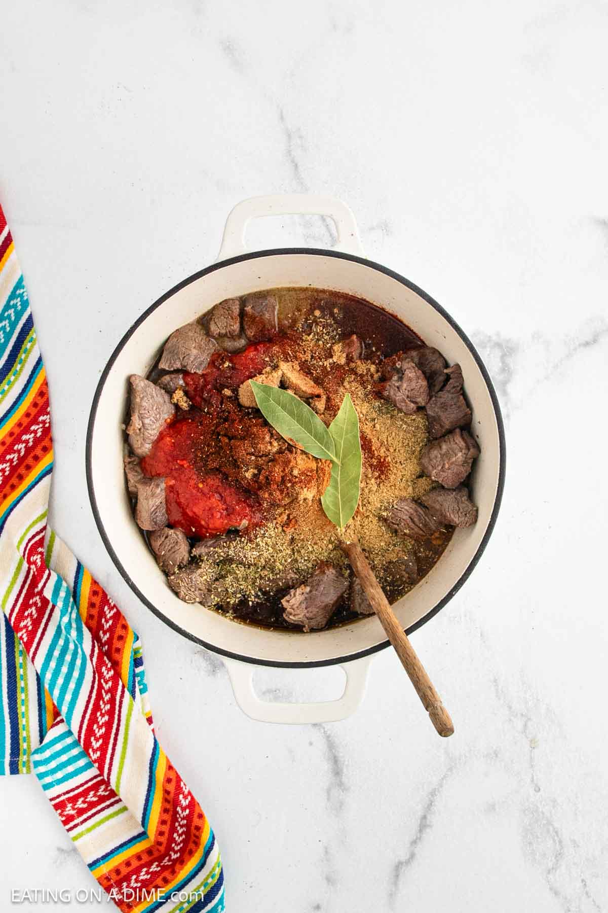 A white pot on a marble surface contains chunks of beef, tomato sauce, spices, and a bay leaf, with a wooden spoon resting inside. A colorful striped cloth is placed to the left of the pot.