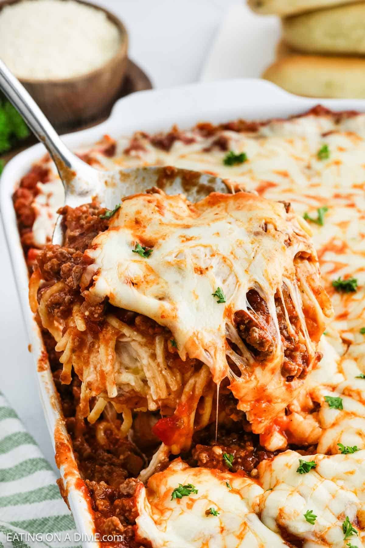 A close-up of a baked Alfredo Spaghetti casserole recipe being served, with melted cheese stretching as a slice is lifted from a white dish. The golden-brown cheese topping is garnished with chopped herbs—perfect for your next viral TikTok!.