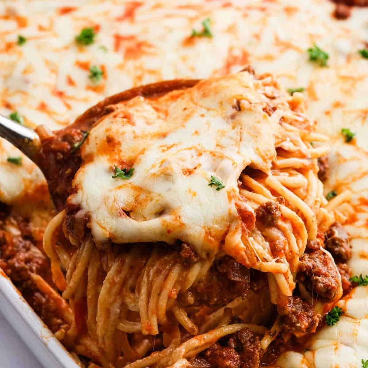 A close-up of a serving of cheesy baked spaghetti with ground beef being lifted from a casserole dish, topped with melted cheese and garnished with chopped parsleyโthis TikTok recipe puts a fun twist on classic Alfredo Spaghetti.
