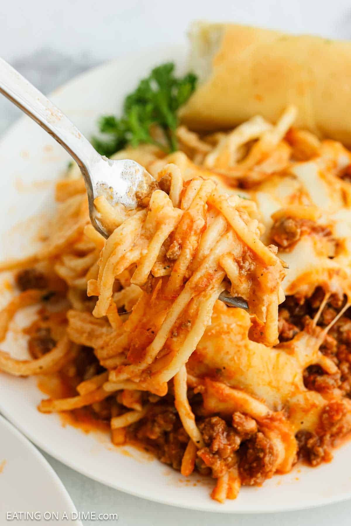 A fork lifts a serving of cheesy baked Alfredo Spaghetti with meat sauce from a white plate; a breadstick and parsley garnish are visible in the background—try this recipe trending on TikTok.