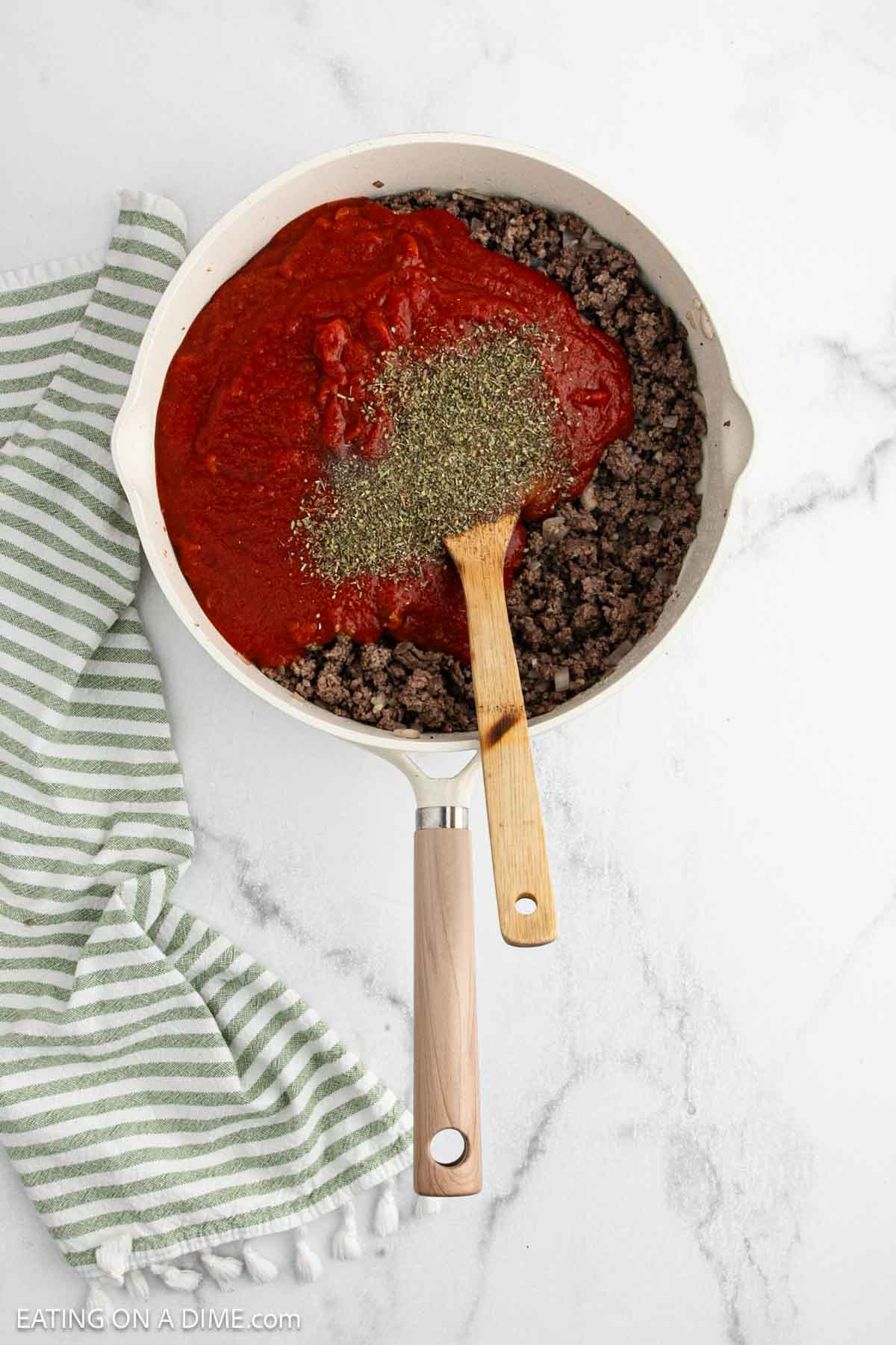 A skillet with cooked ground beef, tomato sauce, and Italian herbs stirred with a wooden spoon—like a savory twist on TikTok pasta. A green and white striped kitchen towel sits beside the pan on a white marble surface.