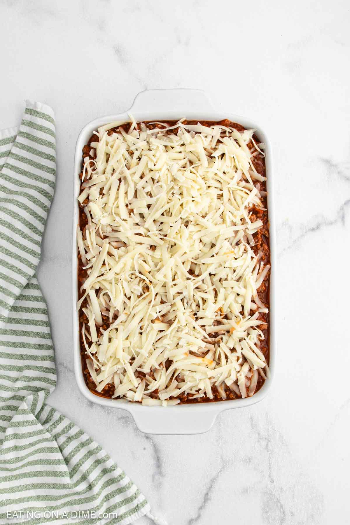 A white baking dish filled with an unbaked Alfredo Spaghetti casserole, topped with shredded cheese, sits on a marble surface next to a green and white striped kitchen towel—perfect for trying that trending TikTok recipe.