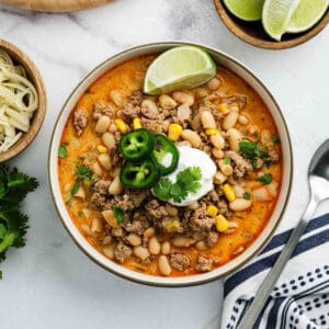 A bowl of White Bean Turkey Chili topped with sour cream, jalapeรฑo slices, cilantro, and a lime wedge. Visible ingredients include ground turkey, white beans, corn, and a creamy broth. A spoon, cheese, lime wedges, and herbs are nearby.