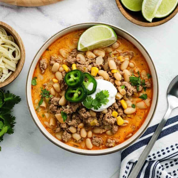 A bowl of White Bean Turkey Chili topped with sour cream, jalapeño slices, cilantro, and a lime wedge. Visible ingredients include ground turkey, white beans, corn, and a creamy broth. A spoon, cheese, lime wedges, and herbs are nearby.