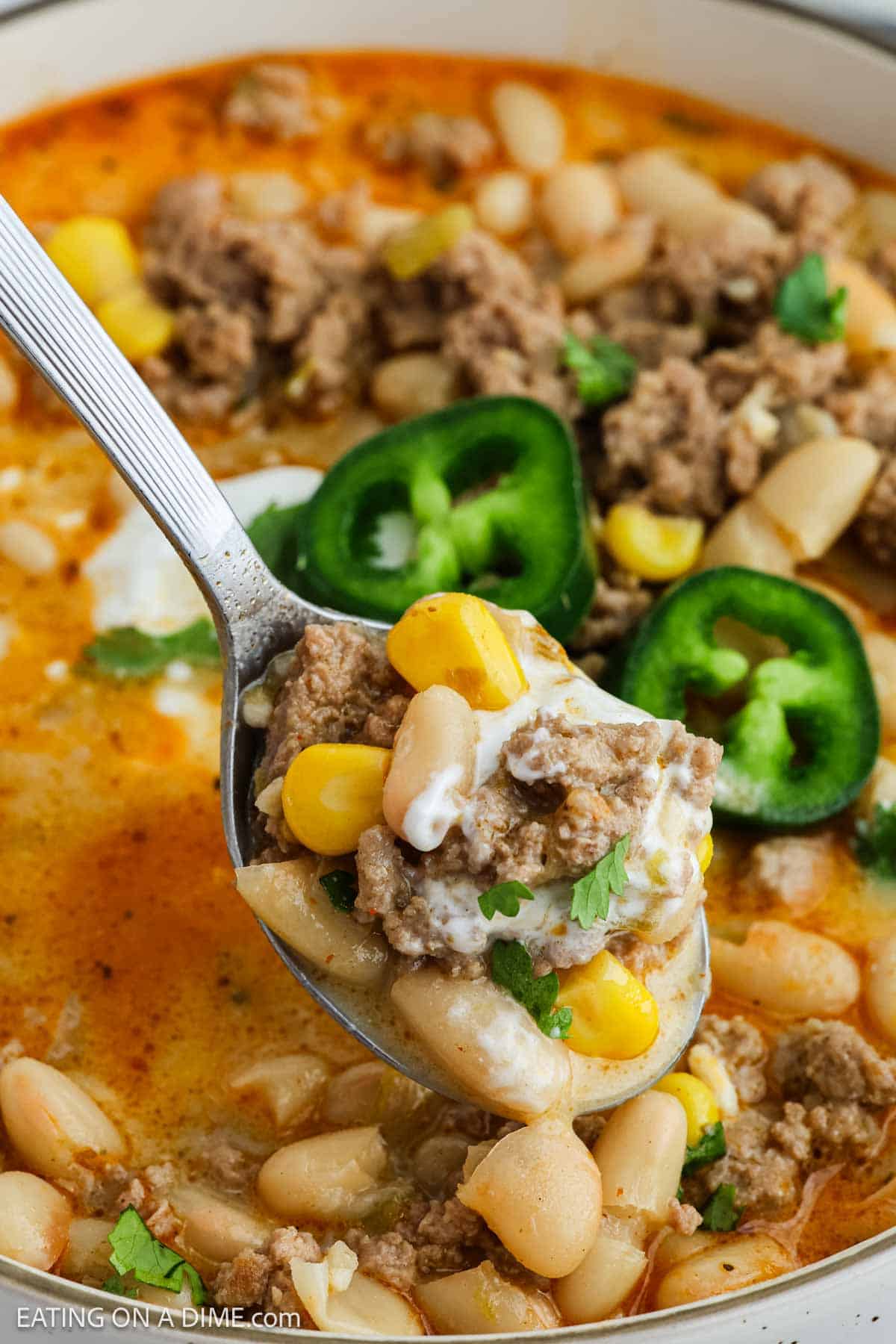 A close-up of a spoonful of White Bean Turkey Chili with ground turkey, white beans, corn, sliced jalapeño, cilantro, and sour cream held over a bowl of the same hearty chili.