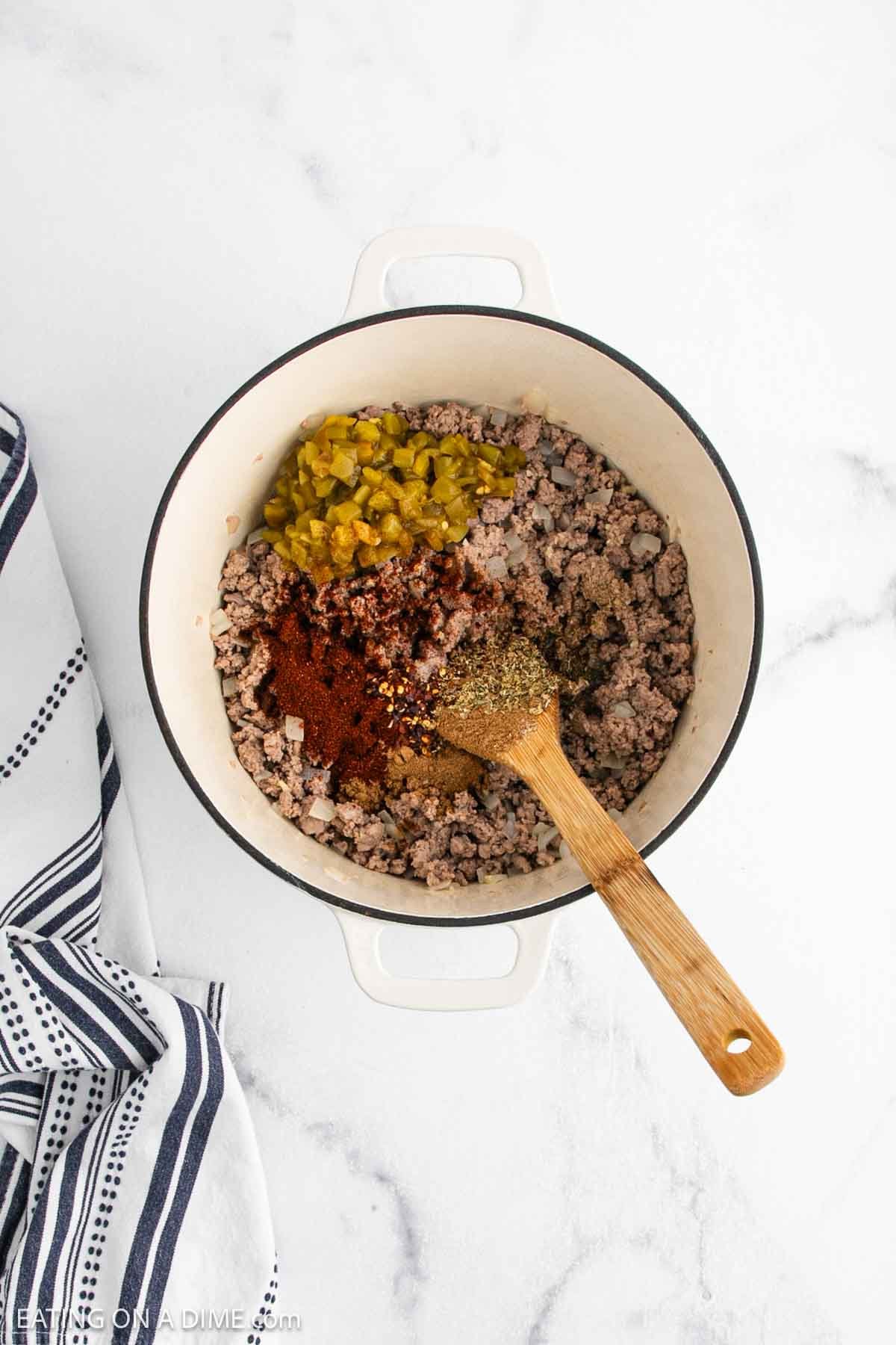 A white pot filled with cooked ground turkey, chopped green chiles, white beans, and various spices for a savory White Bean Turkey Chili. A wooden spoon stirs the mixture as a striped kitchen towel rests on the white countertop.